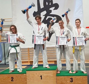 Szymon Bloch z dyplomem i pucharem, ubrany w karate-gi, obok inni sportowcy na podium, Szymon na najwyższym stopniu z numerem 1