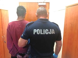 policjant trzyma zatrzymanego, który ma na rekach z tyłu kajdanki, osoby odwrócone plecami