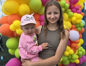 dziewczyna trzymie małe dziecko na rękach, pozuje do zdjęcia, w tle kolorowe balony, słoneczna pogoda
