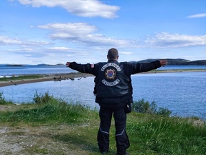 motocyklista stoi z rozłożonymi rękoma, spogląda na jezioro, w tle góry, słonecznie, na niebie nieliczne chmury