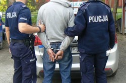 policjanci trzymają osobę z kajdankami na rękach z tyłu, dzień, stoją przed radiowozem