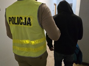 policjant prowadzi mężczyznę, który ma kaptur na głowie, idą korytarzem