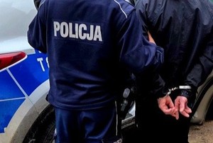 policjant stoi obok zatrzymanego, który ma kajdanki z tylu, dzień, w tle radiowóz policji