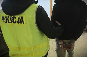 zdjęcie -policjant prowadzi osobę w kajdankach, korytarz budynku