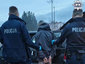 policjanci prowadzą zatrzymanego, pochmurny dzień, parking, w tle samochód