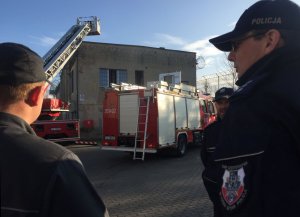 policjant ze strażnikiem więziennym w tle budynek i radiowóz straży pożarnej