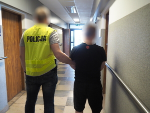 policjant z kamizelce odblaskowej trzyma za ramię zatrzymanego - stoją na korytarzu, tyłem do fotografującego