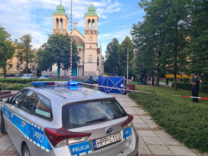 zdjęcie na pierwszym planie przedstawia radiowóz stojący w miejscu zabójstwa w Częstochowie na placu Orląt Lwowskich, na drugim planie rozwinięty parawan oraz grupa policjantów na miejscy zdarzenia