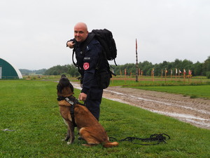 policjant nachyla się nad psem, który siedzi przy jego nodze na trawie