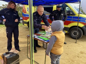 policjant i policjantka stojąc pod namiotem koloru niebeskiego przekazują opaski odblaskowe dzieciom