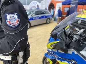 na pierwszym planie naszywka na mundurze policjanta na motocyklu, pawilon profilaktyczny policji w tle i radiowóz