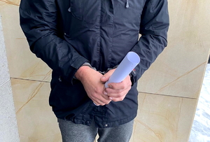 zdjęcie: mężczyzna z kajdankami na rękach ubrany w granatową kurtkę i szare spodnie. W rękach trzyma rulon zwinięty z kartki