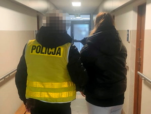 na zdjęciu policjant w kamizelce odblaskowej trzyma za ramię zatrzymaną kobietę na korytarzu komendy