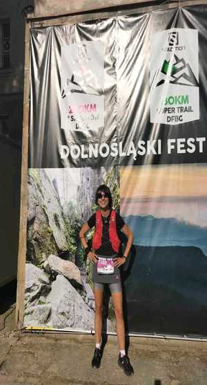 Uśmiechnięta kobieta w stroju sportowym na tle plakatu z napisem &quot;Dolnośląski Festiwal Biegów Górskich&quot;.