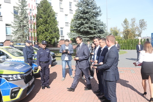 Delegacja kilku mężczyzn idzie w kierunku policjanta, któremu zostaną wręczone oficjalnie kluczyki do nowego radiowozu