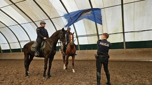 Policjant macha chorągwią przed końmi na których siedzą policjanci