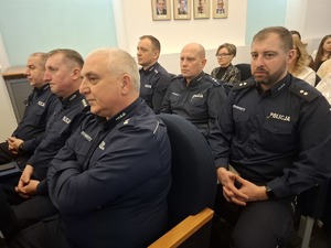 Policjanci w umundurowaniu służbowym siedzą w wyznaczonych miejscach w sali sesyjnej