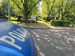 Policyjni jeźdźcy i policyjny przewodnik z psem stoją w parku, w tle napis Policja na karoserii radiowozu