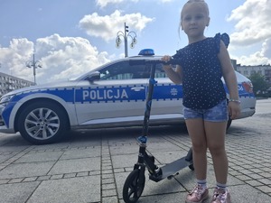 Dziewczynka na hulajnodze pozuje do zdjęcia na tle radiowozu