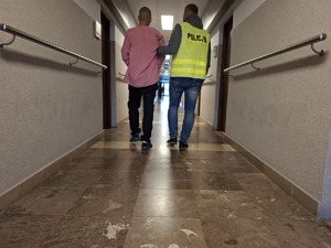 Policjant korytarzem doprowadza zatrzymanego w kajdankach zespolonych