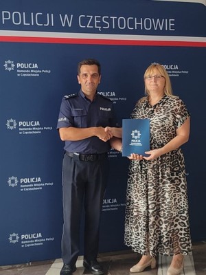 Komendant w mundurze podaje prawą rękę kobiecie i wspólnie pozują do zdjęcia przy ściance z napisem Policja