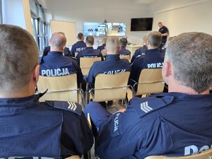 Policjanci siedzą w sali i z ekranu słuchają przemowy policjanta ze szkoły policji online