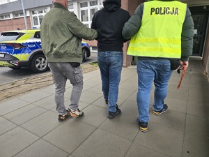 Policjanci doprowadzają zatrzymanego, obok zaparkowany radiowóz
