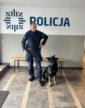 Policjant ze swoim psem stoi przy ścianie z napisem Policja