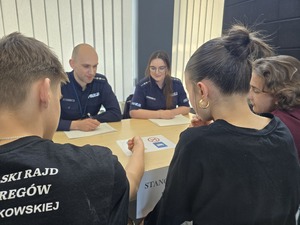 Policjanci egzaminują drużynę, która siedzi przed nimi przy stoliku i odpowiada na zadawane pytania