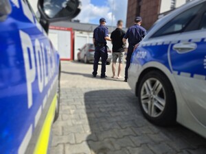 Policjanci doprowadzają zatrzymanego na parkingu samochodowym, gdzie parkują policyjne radiowozy