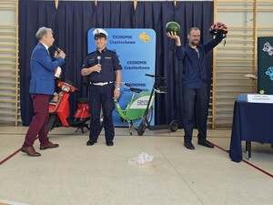 Policjant mówi do mikrofonu podczas akademii szkolnej, obok stoi mężczyzna, który w jednej ręce trzyma kask, a w drugiej arbuza