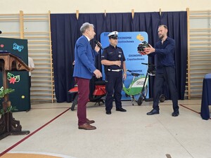 Mężczyzna trzyma w ręce arbuza w kasku ochronnym, obok stoi policjant i dyrektor szkoły