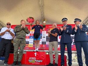 kobieta stoi na podium, trzymając puchar i kwiaty w górze, obok stoją policjanci w mundurach i mężczyzna w mundurze wojskowym i biją brawo
