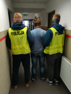 Zdjęcie przedstawia dwóch nieumundurowanych policjantów w kamizelkach odblaskowych z napisem POLICJA, prowadzących zatrzymanego korytarzem KMP Dąbrowa Górnicza. Zatrzymany ma założone kajdanki na ręce trzymane z przodu.