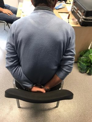 Zdjęcie przedstawia zatrzymanego siedzącego z boku biurka, który jest przesłuchiwany przez policjanta pionu kryminalnego. Zatrzymany ma założone kajdanki na ręce trzymane z tyłu.