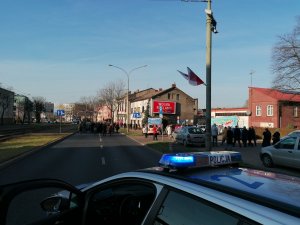 Zdjęcia przedstawiające na pierwszym planie oznakowany radiowóz policyjny używający niebieskich sygnałów świetlnych oraz w tle uczestników przemarszu ulicami Dąbrowy Górniczej.