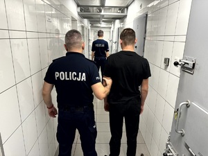 Dwóch umundurowanych policjantów prowadzących korytarzem zatrzymanego.