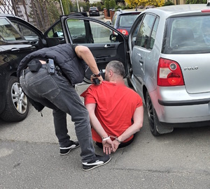 Zatrzymany mężczyzna z kajdankami na rekach trzymanych z tyłu, sprawdzany przez nieumundurowanego policjanta.