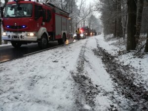 Leśna droga, a na niej wóz strażacki na pierwszym planie, za nim kolejny podobny i na końcu radiowzóz. Widoczne w śniegu ślady samochodu, który zjechał z asfaltu i koleiny prowadza w stronę pobocza.