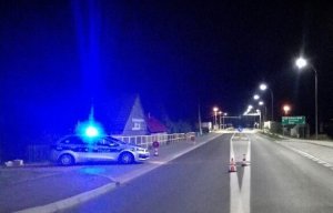Po lewej zaparkowany policyjny radiowóz z włączonymi sygnałami świetlnymi. Na wprost most i pusta nocna droga.