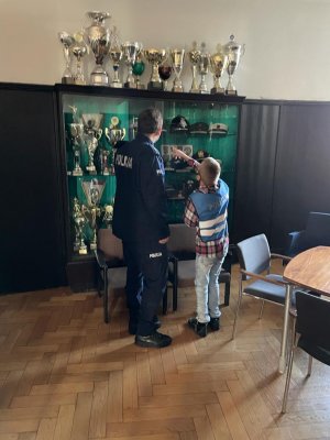 Policjant pokazuje chłopcu puchary i czapki policji z rożnych krajów - wszystko wyeksponowane w gablocie.