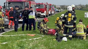 Na zdjęciu widzimy grupę strażaków, ratowników medycznych i policjantów skupionych na ratowaniu rannych ułożonych na trawniku.