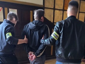 Na zdjęciu dwóch policjantów nieumundurowanych prowadzi zatrzymanego w kajdankach.