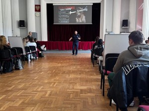 Na dużej sali siedzi grupa młodzieży podczas wykładów.. Na środku sali stoi umundurowany policjant, który prowadzi wykład.