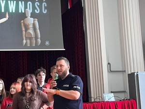 Na dużej sali siedzi grupa młodzieży podczas wykładów. Policjant z mikrofonem w ręku prowadzi wykład.