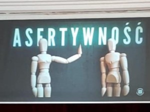 Na dużej sali siedzi grupa młodzieży podczas wykładów. Policjant z mikrofonem w ręku prowadzi wykład stojąc przy scenie wraz z policjantką.