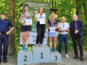 Trzy policjantki stojące na zwycięskim podium. Obok dwóch umundurowanych policjantów i dwóch mężczyzn w cywilnych ubraniach.