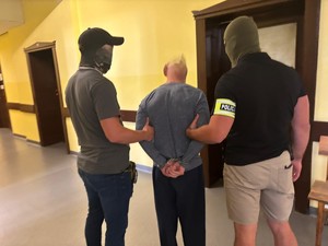 Na korytarzy stoi zatrzymany, którego pilnują nieumundurowani policjanci.