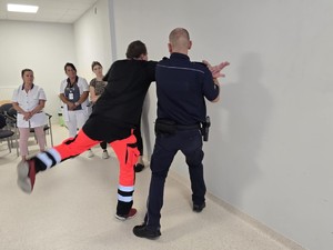 Policjant i mężczyzna w stroju ratownika ćwiczą chwyty obronne przy ścianie.