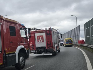 Na jezdni stoi Straż Pożarna i radiowozy Policji. Pomiędzy nimi przemieszczają się funkcjonariusze obsługujący wypadek drogowy.. W tle na poboczu stoi czerwony parawan z napisem STRAŻ.